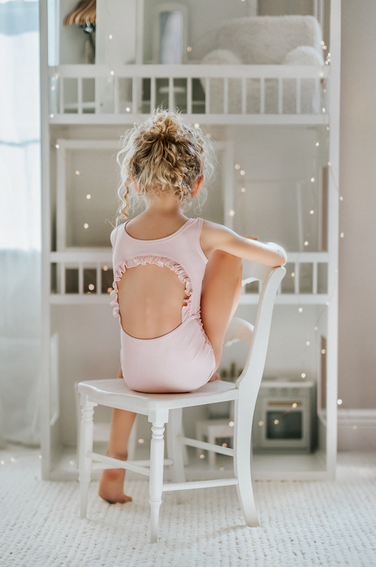 A young girl with curly hair is sitting on a white chair in a room adorned with string lights. She is dressed in a pink Ruffle Back Leotard, which is crafted from a poly/spandex blend. There is a back cutout lined with ruffles.