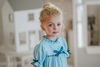 A young girl is wearing The Original Clara Nutcracker Gown in Robbins Egg Blue, featuring blue ruffles and a navy bow. The buttery soft fabric is cozy and sensory friendly.  The puff sleeves with ribbon detail and the ruffles at the hem add a traditional classic look. 