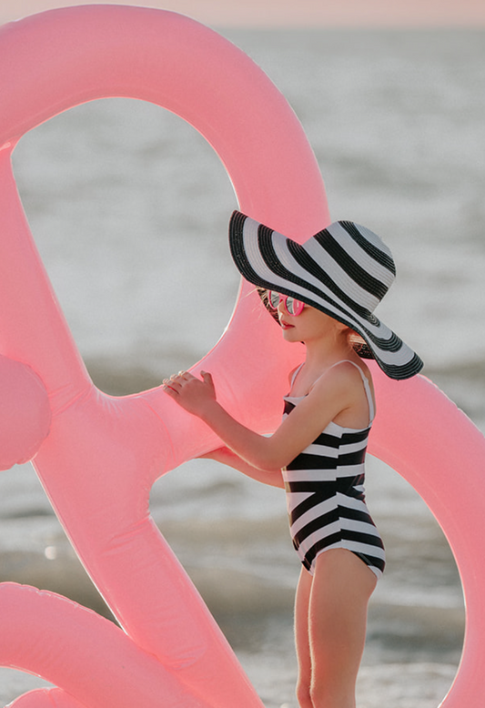 A young girl is wearing a black and white stripped one piece swim suit.  The white spaghetti straps are adjustable. The black and white pattern is in a chevron style.  A large floppy brimmed beach hat in matching black and white stripe is on her head. 