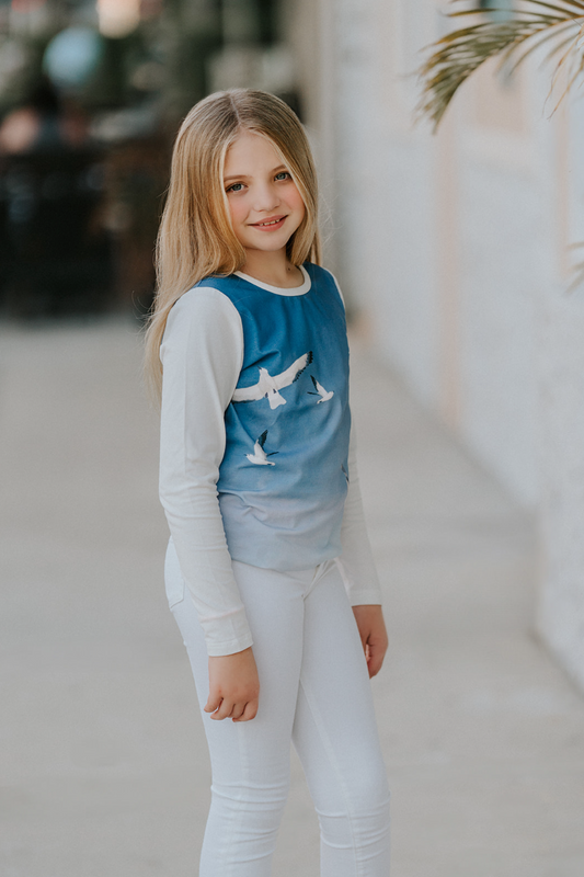 A young girl stands outdoors, smiling at the camera. She has long blonde hair and is wearing a stylish Seagull Soundtrack Shirt with a blue front panel featuring birds in flight, crafted from soft cotton jersey knit, paired with white leggings. The background is slightly blurred with a touch of greenery.