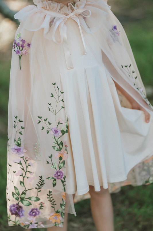 A young girl is dressed in a spring floral tutu dress. Lavender velvet bodice with short sleeves and fluffly layers of ivory tulle. The skirt is embroidered with peach, purple, pink flowers and green vines. A matching ivory cape with floral pattern is tied around her shoulders. 