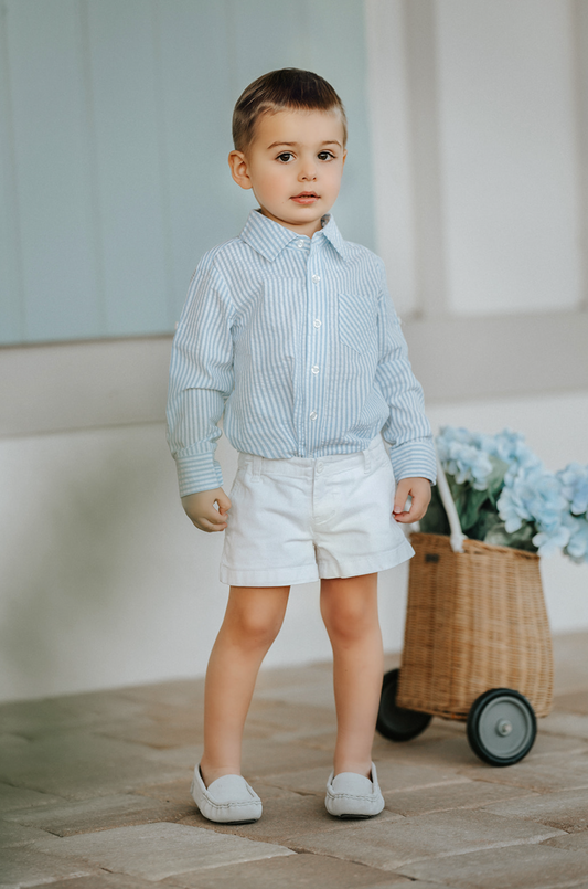 A young boy wears a long sleeve button up collared shirt. A light blue and white vertical seersucker shirt with a pocket on the left chest. 