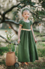 A young girl is wearing a Renaissance Princess Gown Guinevere Historical Maxi Dress in green with gold accents. Her yellow and green headband complements her chic ensemble.  The neckline and puff sleeves have gold trim.  The middle panel of the maxi dress is has matching earthy green lace.