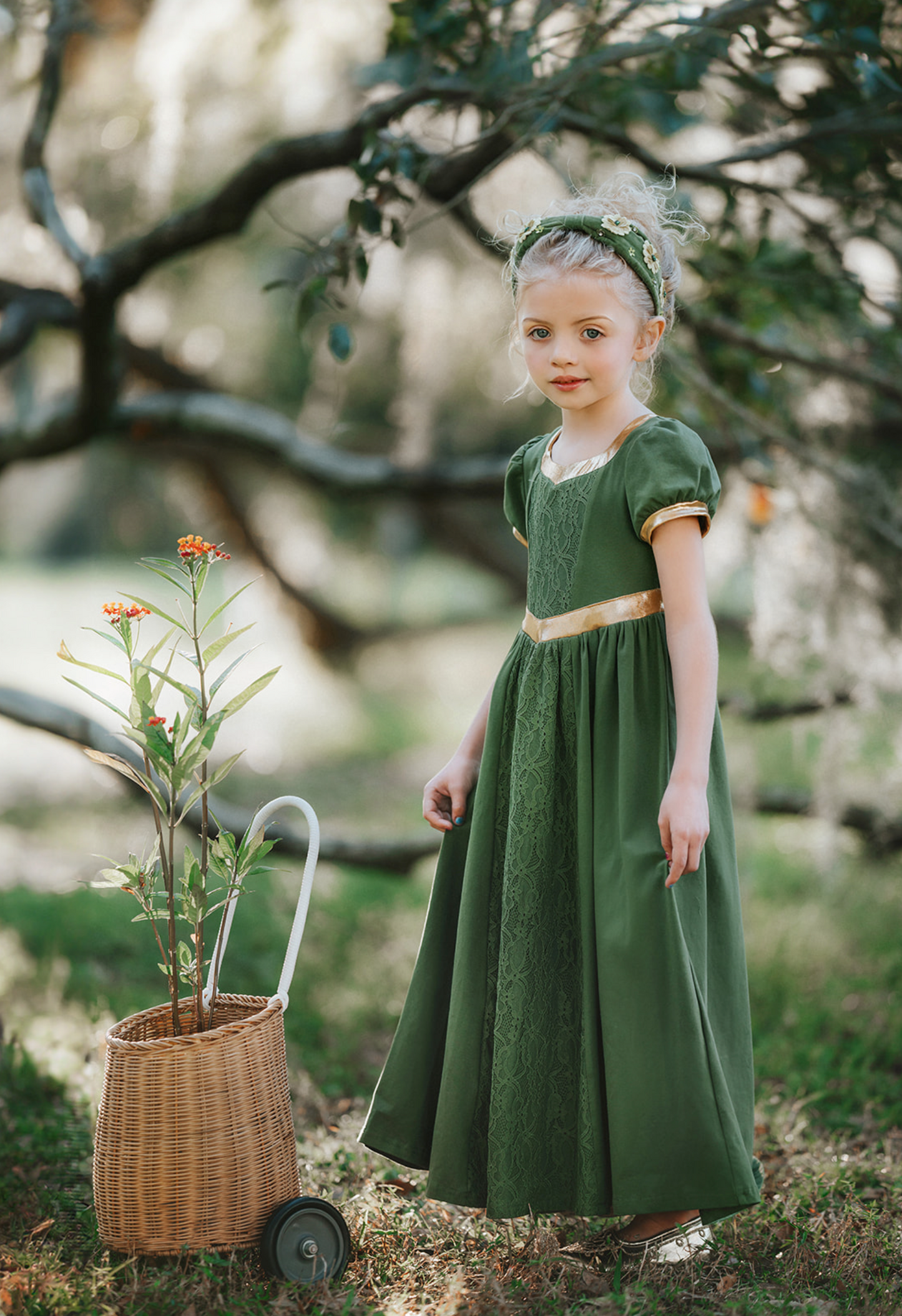 A young girl stands outdoors, surrounded by trees, wearing a Renaissance Princess Gown Guinevere Historical Maxi Dress in green with gold accents. Her leafy headband complements her chic ensemble, and she holds onto a small wicker basket on wheels with a potted flowering plant inside. The scenery is lush and green.