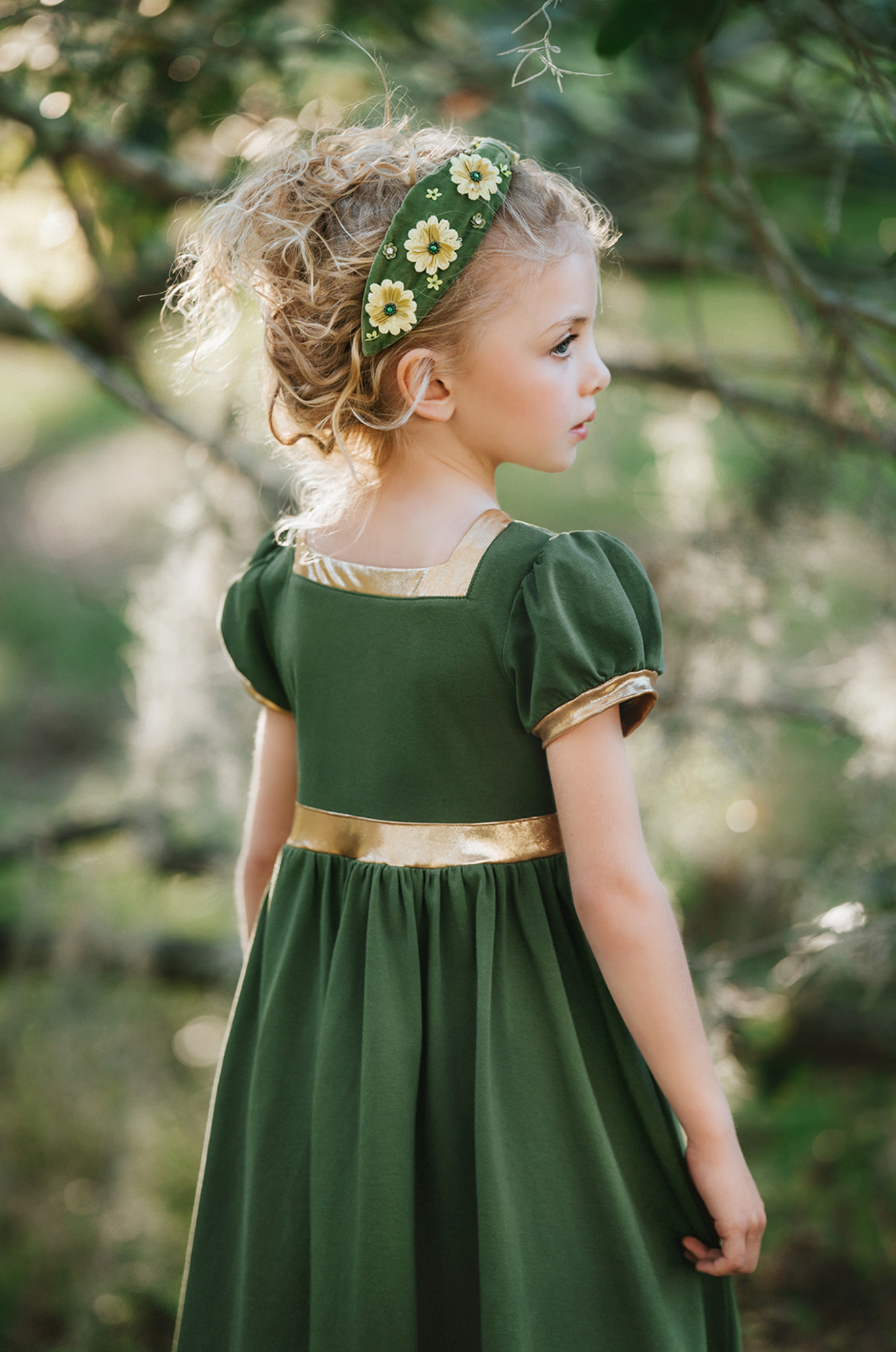 A young girl stands outdoors, surrounded by trees, wearing a Renaissance Princess Gown Guinevere Historical Maxi Dress in green with gold accents. Her leafy headband complements her chic ensemble, and she holds onto a small wicker basket on wheels with a potted flowering plant inside. The scenery is lush and green.