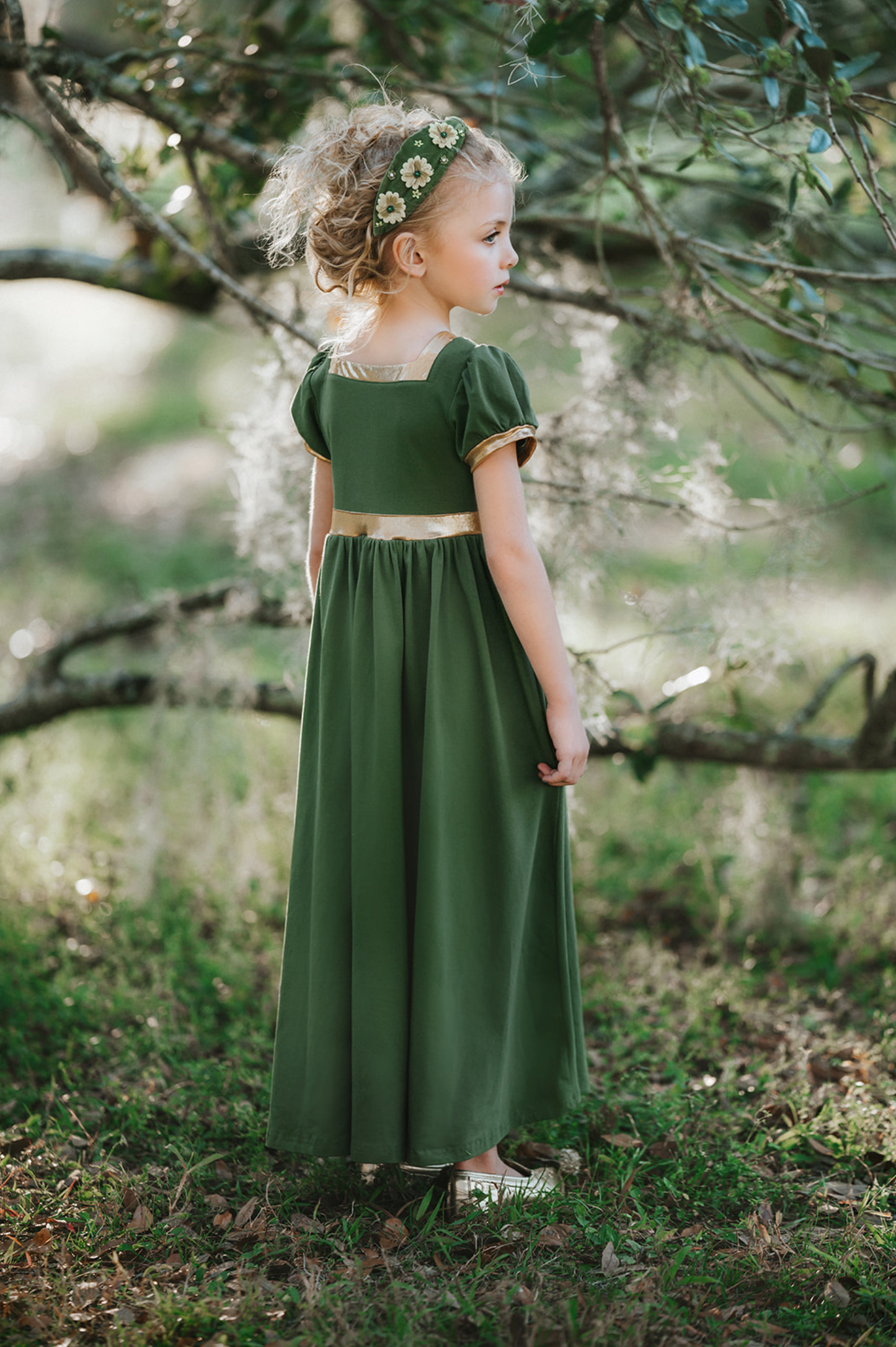 A young girl stands outdoors, surrounded by trees, wearing a Renaissance Princess Gown Guinevere Historical Maxi Dress in green with gold accents. Her leafy headband complements her chic ensemble, and she holds onto a small wicker basket on wheels with a potted flowering plant inside. The scenery is lush and green.