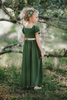 A young girl stands outdoors, surrounded by trees, wearing a Renaissance Princess Gown Guinevere Historical Maxi Dress in green with gold accents. Her leafy headband complements her chic ensemble, and she holds onto a small wicker basket on wheels with a potted flowering plant inside. The scenery is lush and green.