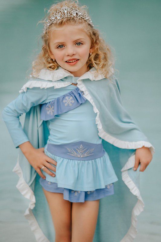 A young girl is wearing a light blue terry cloth and cotton swim cover up.  The cape has a hood and white ruffle trim along all the edges.  There is a snap closure at the neck. 