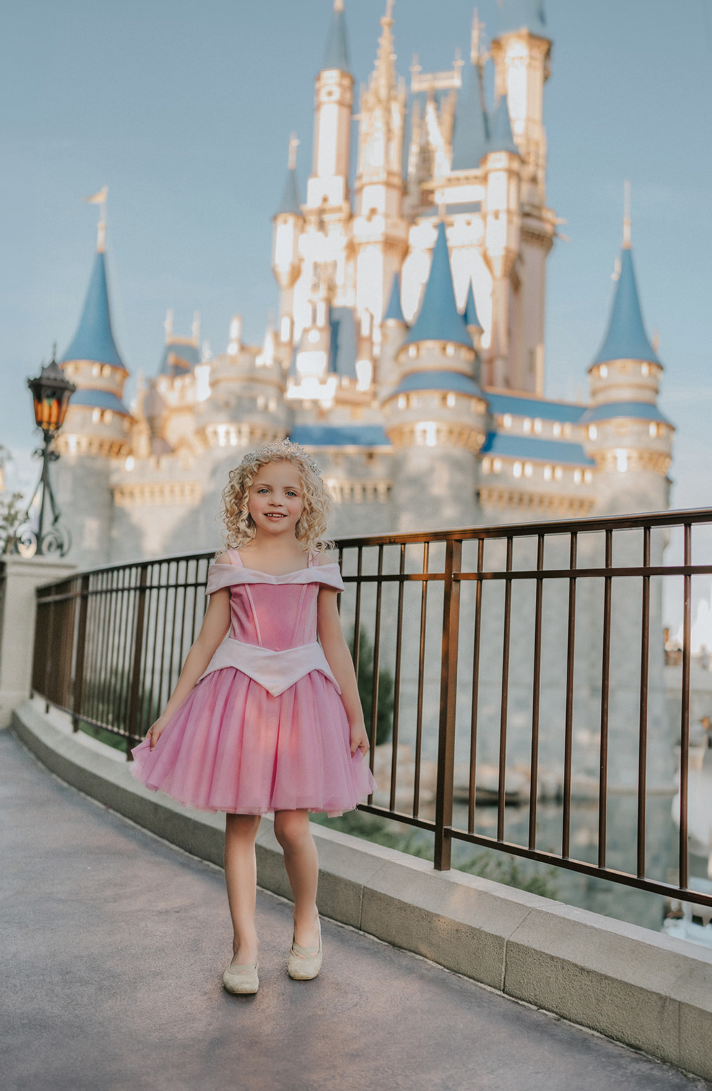 A young girl in an exquisite Portrait Petite Sleeping Beauty twirl length gown poses in a castle setting.  The pink stretch velvet gown has an off the shoulder neckline with two straps.   The pink tulle skirt is full and has a cotton liner for comfort.