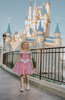 A young girl in an exquisite Portrait Petite Sleeping Beauty twirl length gown poses in a castle setting.  The pink stretch velvet gown has an off the shoulder neckline with two straps.   The pink tulle skirt is full and has a cotton liner for comfort.