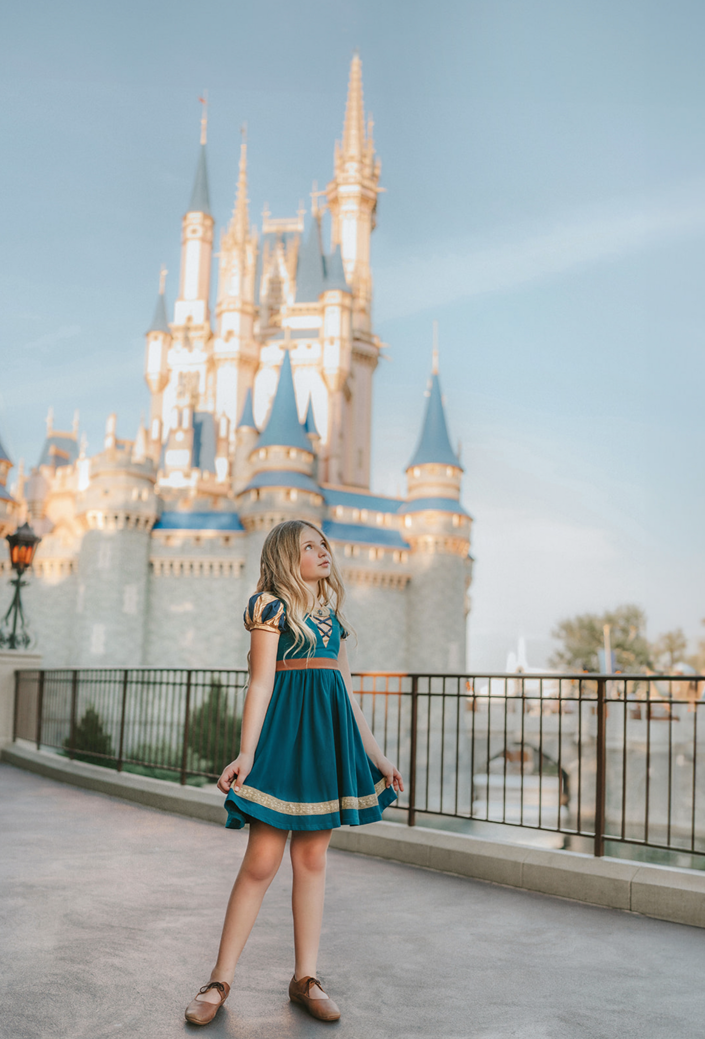 A young girl is wearing a dark teal cotton twirl dress.  The puff sleeves have metallic gold.  Metallic gold lines the neck with a dark teal jewel.  A intricate gold ribbon runs along the hem. 