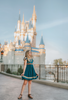 A young girl is wearing a dark teal cotton twirl dress.  The puff sleeves have metallic gold.  Metallic gold lines the neck with a dark teal jewel.  A intricate gold ribbon runs along the hem. 