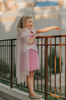 A young girl with curly blonde hair, wearing a tiara and the Portrait Petites Sleeping Beauty Twirl Length Gown adorned with heirloom-quality lace and a sheer white cape, smiles and points while standing near a railing. The background reveals an elegantly blurred stone building with ornate detailing.