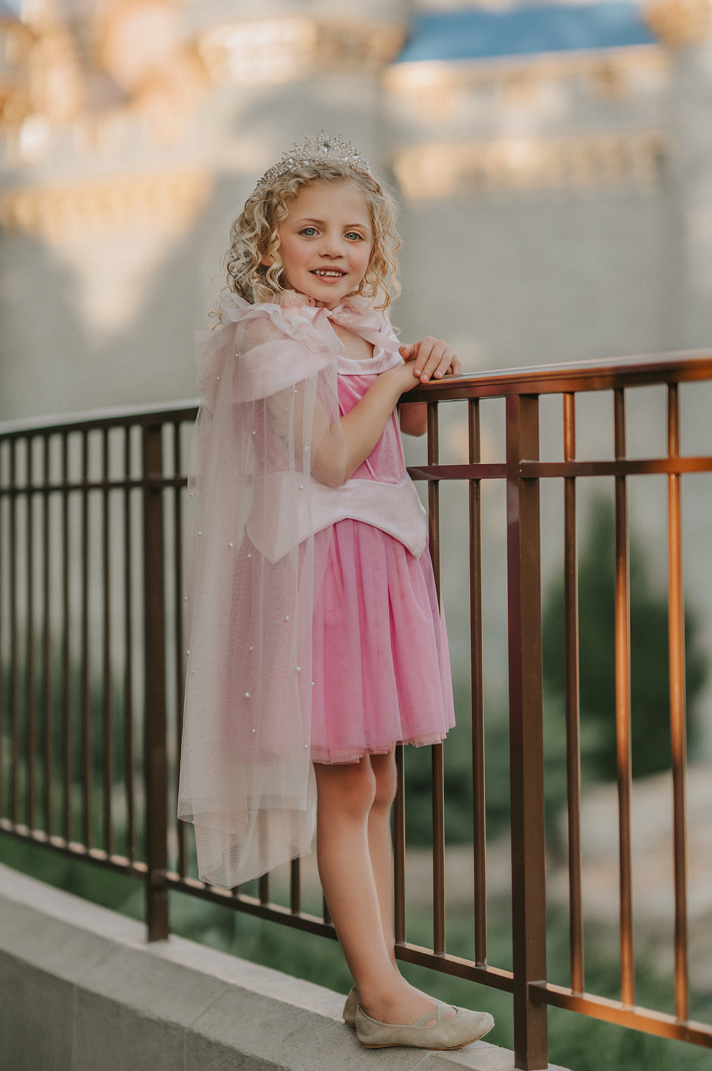 A young girl in an exquisite Portrait Petite Sleeping Beauty twirl length gown poses in a castle setting.  The pink stretch velvet gown has an off the shoulder neckline with two straps.   The pink tulle skirt is full and has a cotton liner for comfort.