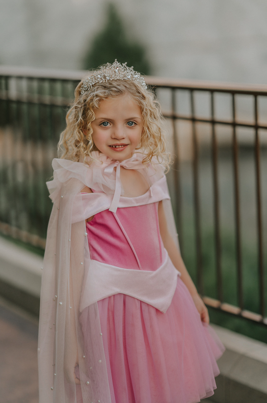 A young girl wears a soft tulle light pink cape tied at the shoulders with a satin ribbon.  The cape has beautiful pearls placed all over and a ruffle collar. 