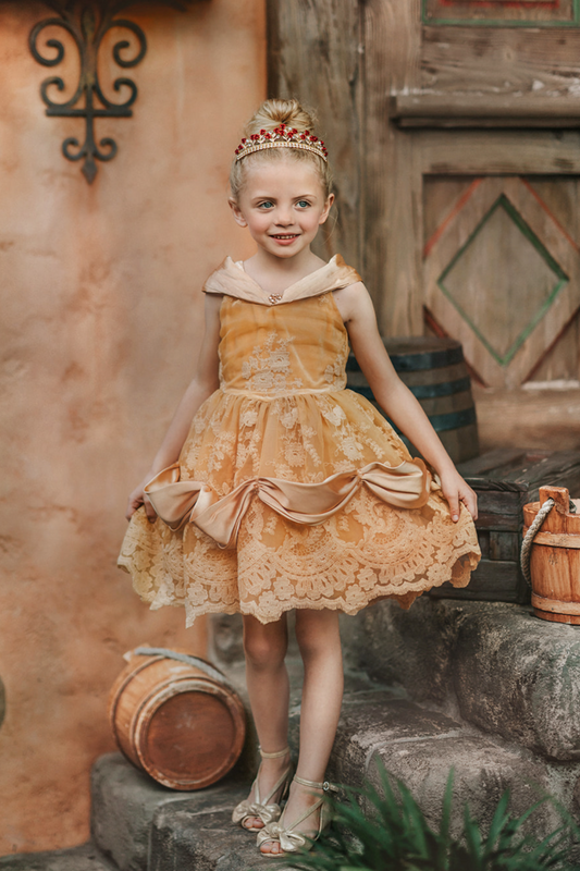 A young girl with blonde hair stands outdoors, wearing the Portrait Petite Red Rose Princess twirl length gown, an intricately designed gold princess dress adorned with floral lace patterns. The satin off shoulder neckline is adorned with a gold jewel. 