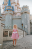 A young girl in an exquisite Portrait Petite Sleeping Beauty twirl length gown poses in a castle setting.  The pink stretch velvet gown has an off the shoulder neckline with two straps.   The pink tulle skirt is full and has a cotton liner for comfort.