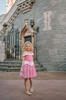 A young girl in an exquisite Portrait Petite Sleeping Beauty twirl length gown poses in a castle setting.  The pink stretch velvet gown has an off the shoulder neckline with two straps.   The pink tulle skirt is full and has a cotton liner for comfort.