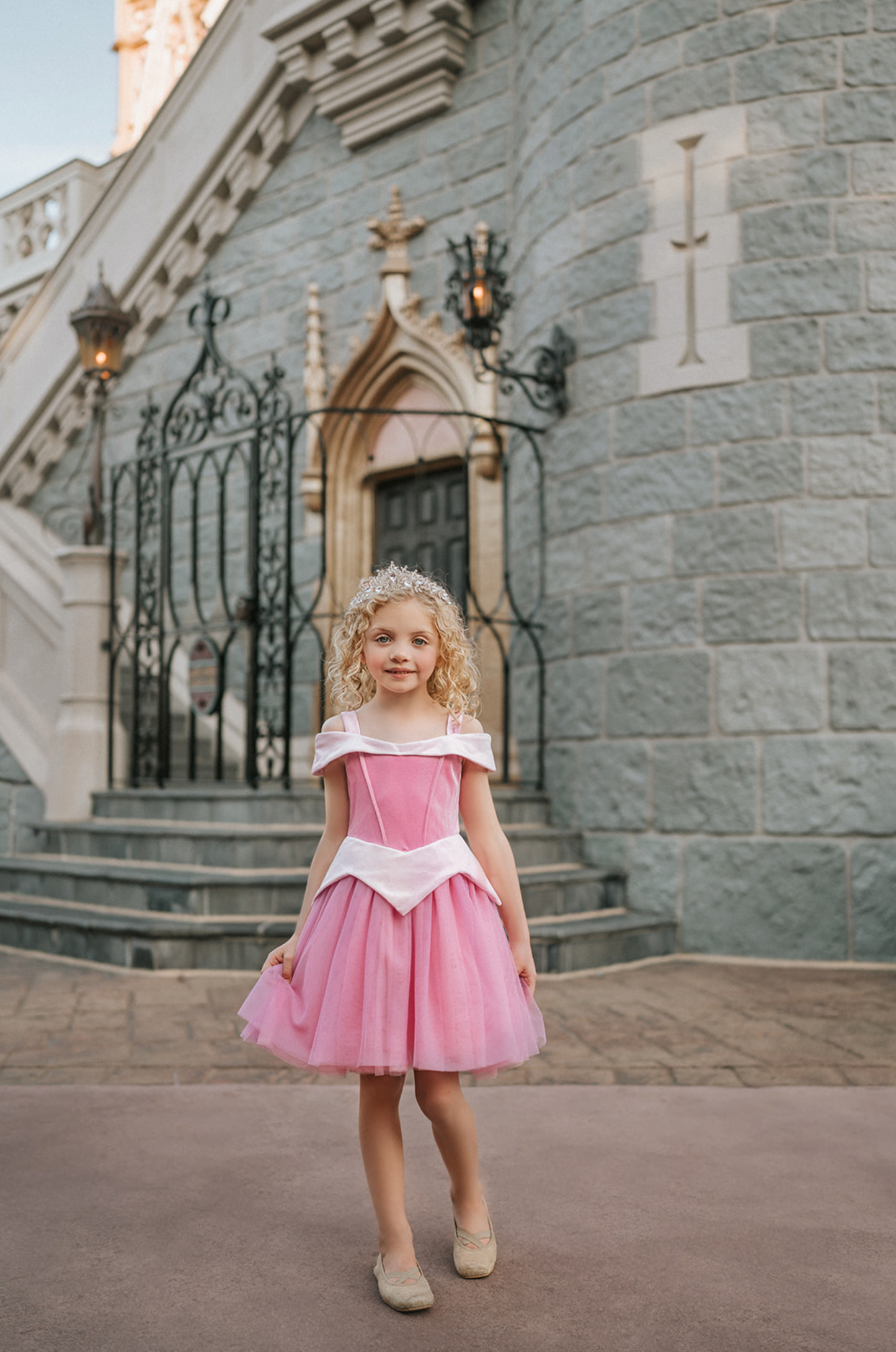 A young girl in an exquisite Portrait Petite Sleeping Beauty twirl length gown poses in a castle setting.  The pink stretch velvet gown has an off the shoulder neckline with two straps.   The pink tulle skirt is full and has a cotton liner for comfort.