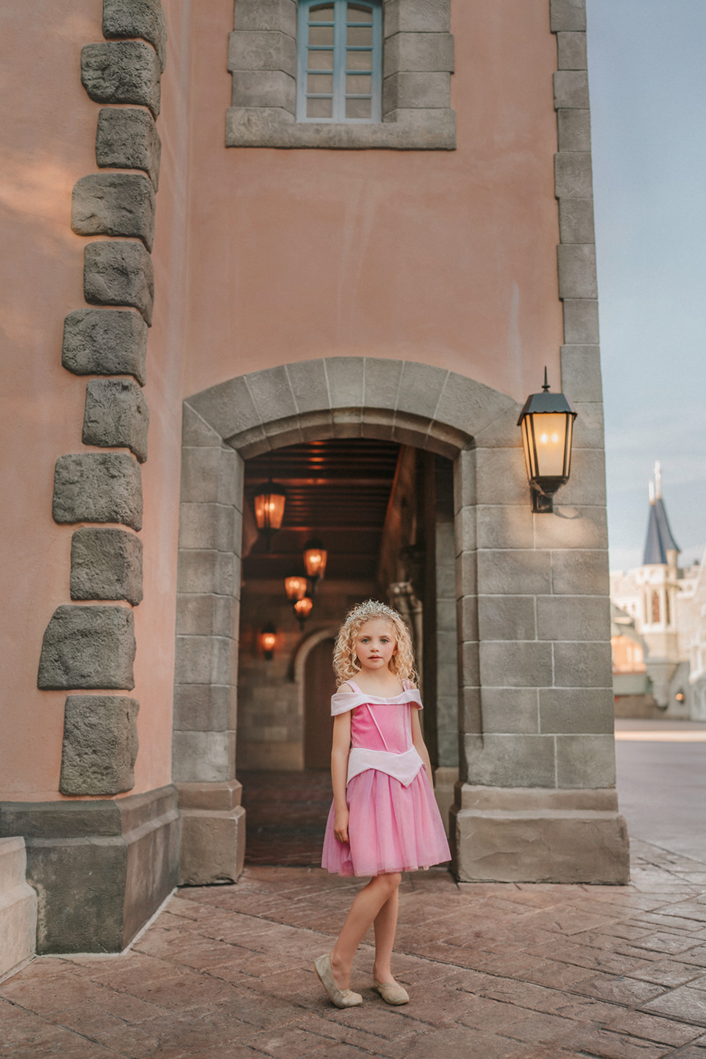 A young girl in an exquisite Portrait Petite Sleeping Beauty twirl length gown poses in a castle setting.  The pink stretch velvet gown has an off the shoulder neckline with two straps.   The pink tulle skirt is full and has a cotton liner for comfort.