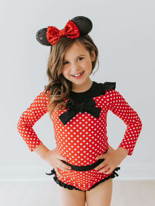 A young girl is wearing our Miss Mouse  Rashguard Swim set.  This 2pc swim has SPF 50.  The top is red with white polka dots.  There is a large black bow in the center and small ruffles on the shoulder.  The bottoms have black ruffles along the edge. 