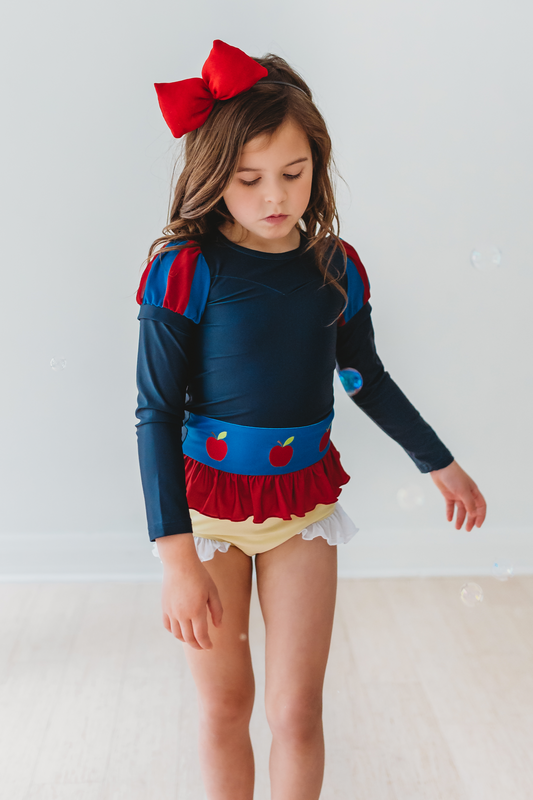 A young girl is wearing the Snow White Rashguard Swim Set.  The long sleeves and spf 50 provide protection from the sun.  The navy blue top has blue and red puff adornments and apples embroidered on a blue band with red ruffle at the hem.  The light yellow bottoms have a white ruffle on the leg. 