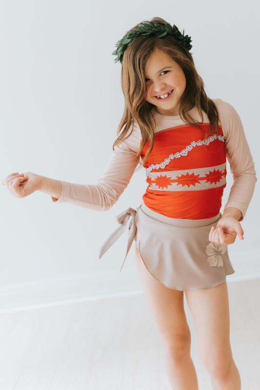 A young girl is wearing a bright red swim top with flower and pattern band.  The tan-grey swimsuit bottom is adorned with a tan hibiscus flower—it's the Polynesian Princess Rashguard Swim Set made from SPF 50 material. 