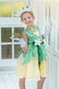 A young girl poses in the Frog Princess Twirl Dress . She wears a green and yellow dress with a magnolia accent at the waist. The light green top and leaf shaped skirt are overlayed on a light yellow skirt with white eyelet trim at the bottom.  The dress is sleeveless and a comfortable cotton. 