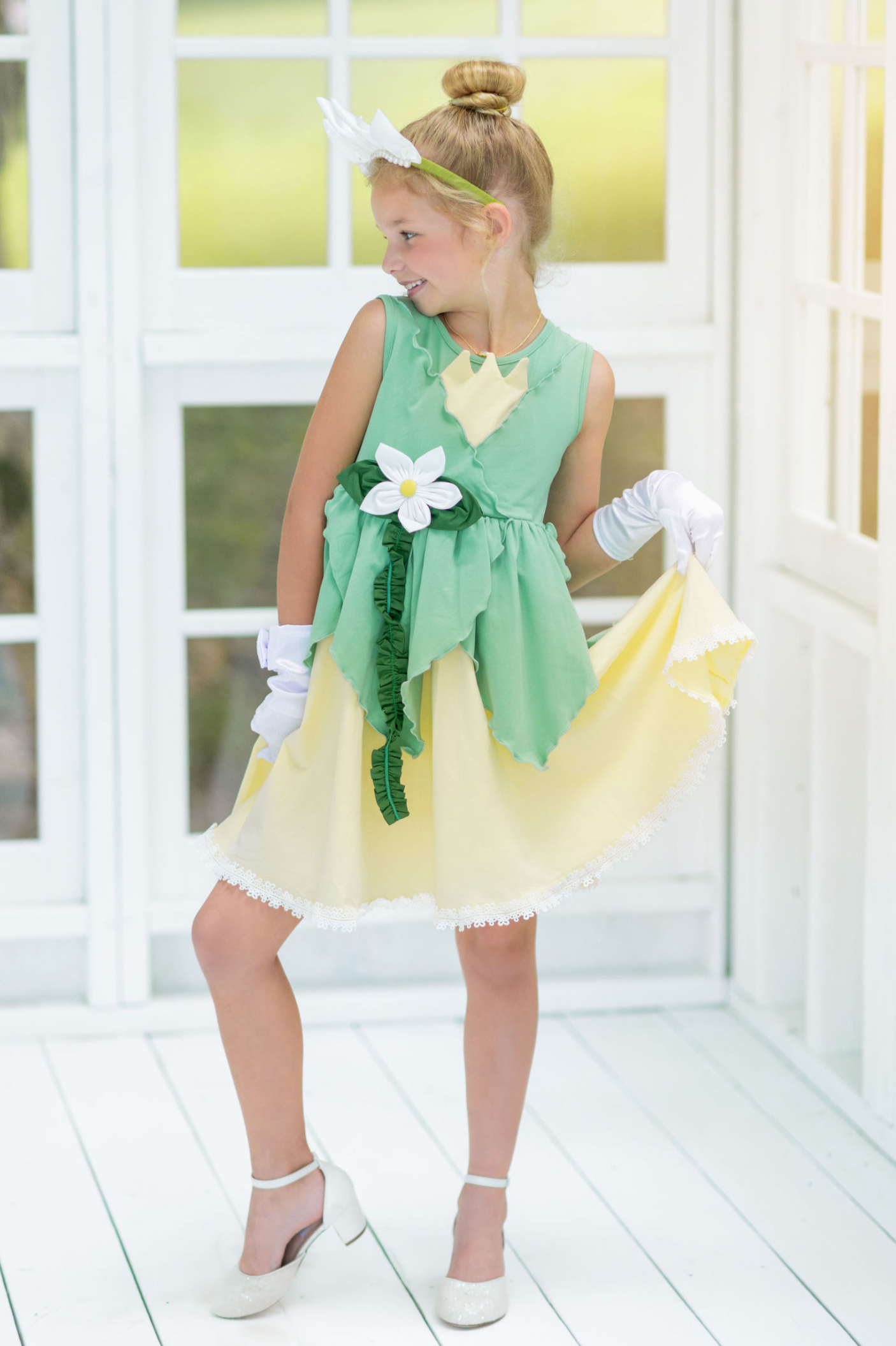 A young girl poses in the Frog Princess Twirl Dress . She wears a green and yellow dress with a magnolia accent at the waist. The light green top and leaf shaped skirt are overlayed on a light yellow skirt with white eyelet trim at the bottom.  The dress is sleeveless and a comfortable cotton. 