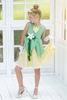 A young girl poses in the Frog Princess Twirl Dress . She wears a green and yellow dress with a magnolia accent at the waist. The light green top and leaf shaped skirt are overlayed on a light yellow skirt with white eyelet trim at the bottom.  The dress is sleeveless and a comfortable cotton. 