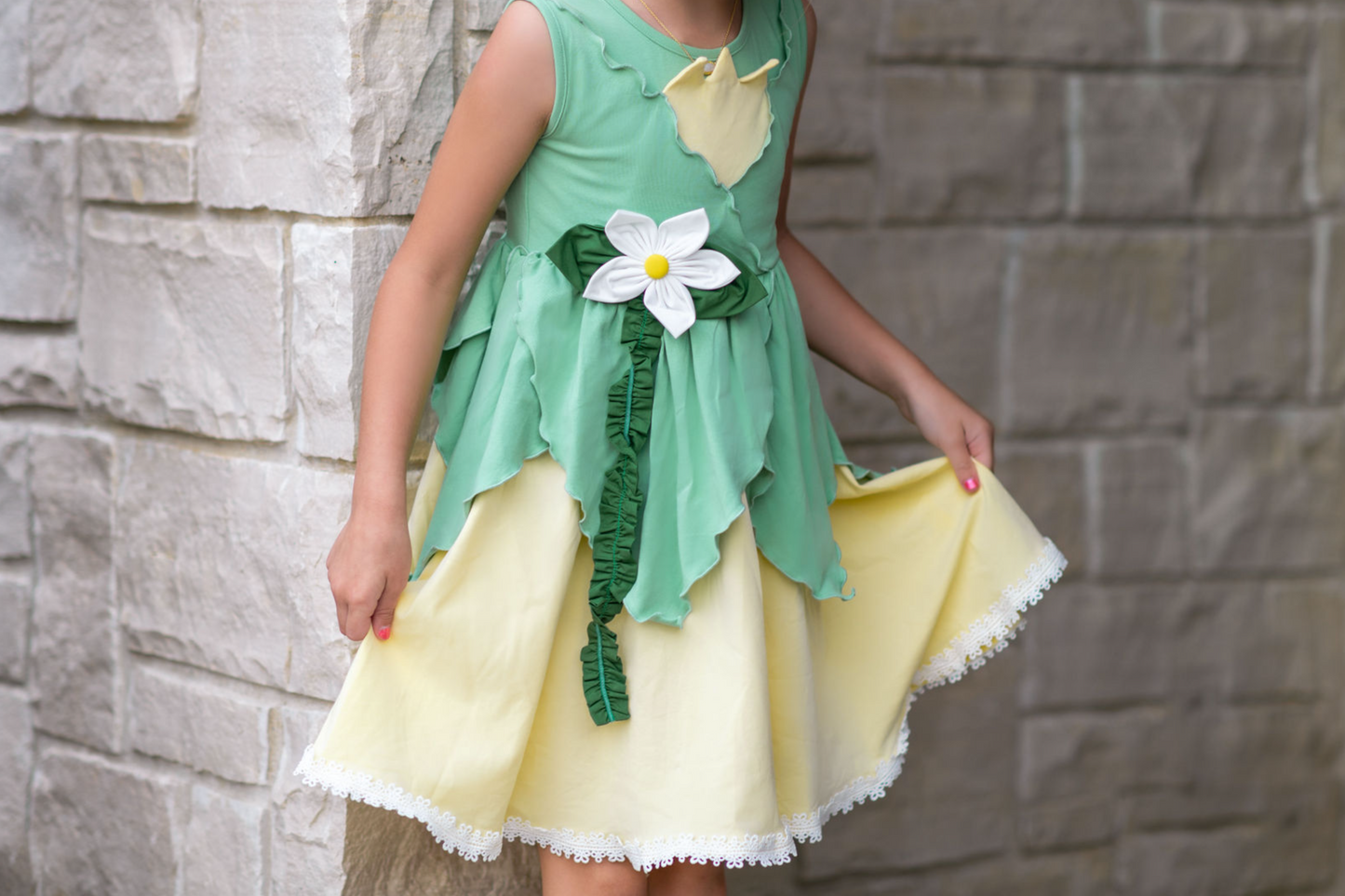 A young girl poses in the Frog Princess Twirl Dress . She wears a green and yellow dress with a magnolia accent at the waist. The light green top and leaf shaped skirt are overlayed on a light yellow skirt with white eyelet trim at the bottom.  The dress is sleeveless and a comfortable cotton. 