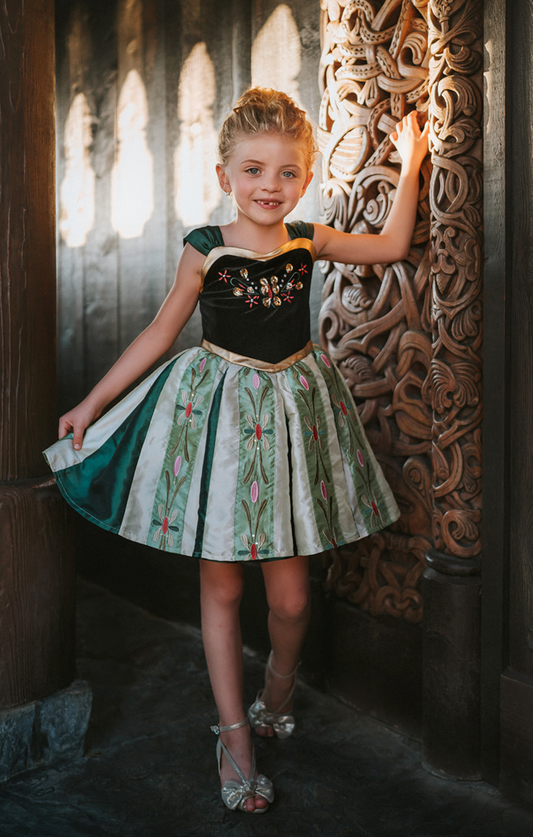 A young girl with blonde hair wearing the Portrait Petites Coronation Ice Princess Twirl Length Gown adorned with custom embroidery in green and black. The black stretch velvet bodice is adorned with golden jewels and flower embroidery. The skirt is shades of green with pink and green embroidered designs. 