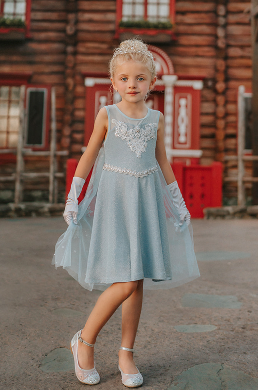 A young girl is dressed in the sparkly light blue Portrait Petite Ice Queen Gown in twirl length adorned with intricate custom  lace at the neckline with rhinestones. A light blue cape with silver sparkle snowflakes is buttoned to the back of the gown. 