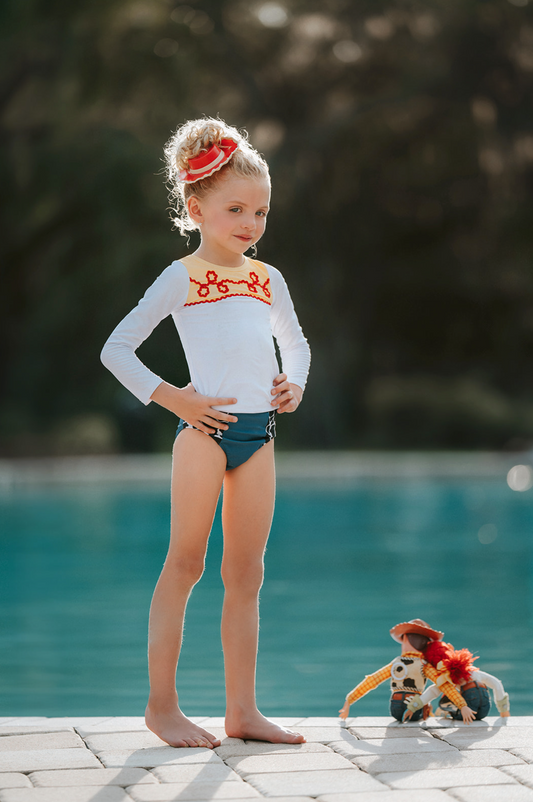 A young girl is wearing a 2 piece long sleeve rash guard swim set.  The top is white with yellow on the upper chest.  Across the yellow is red scrolling rick rack.  The bottoms are a faux denim with black and white cow print panels. 