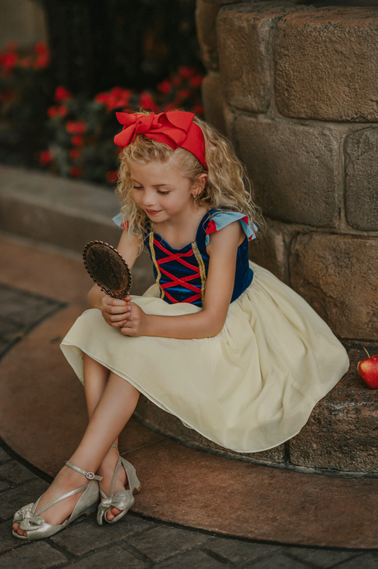 A young girl is wearing the Portrait Petite Twirl Length Snow White Gown. The gown is velvet blue and a light yellow skirt with a red ribbon criss cross accent on the bodice. The dress is sleeveless with a red and blue flutter ruffle on the top. 