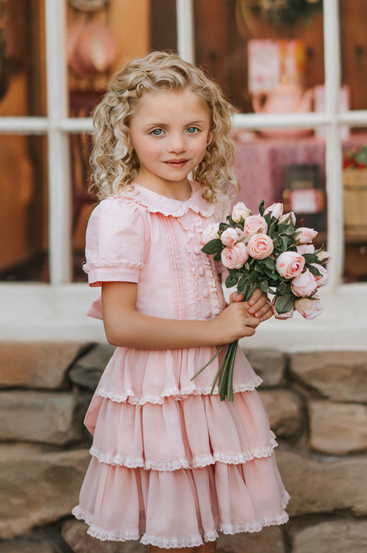 A young girl is wearing our Plaza Girl Pretty Pink Dress.  The beautiful light pink dress has lace details on the peter pan collar and puff sleeves.  There are pink buttons and lace down the front.  The skirt is 3 layers of ruffles that twirl  perfectly.  There is a tie bow in the back. 