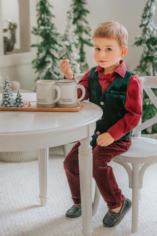 A young boy sits with his hot cocoa. He is wearing an evergreen velvet vest with three flat gold buttons.  There is a small pocket on the left chest.  The back has an adjustable buckle to that tightens.  