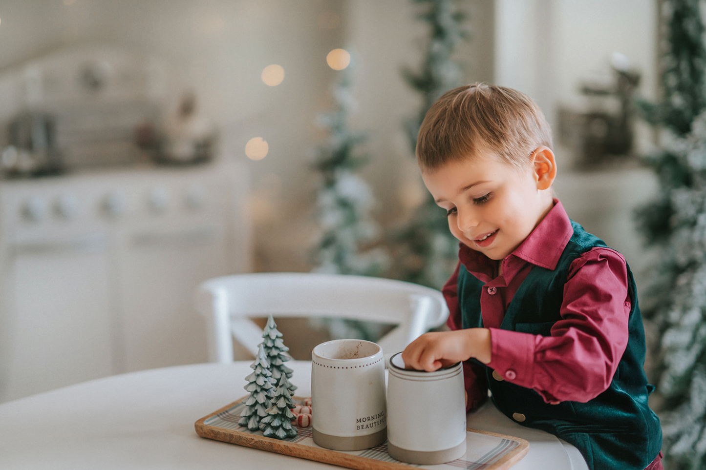 A young boy sits with his hot cocoa. He is wearing an evergreen velvet vest with three flat gold buttons.  There is a small pocket on the left chest.  The back has an adjustable buckle to that tightens.  