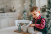 A young boy sits with his hot cocoa. He is wearing an evergreen velvet vest with three flat gold buttons.  There is a small pocket on the left chest.  The back has an adjustable buckle to that tightens.  