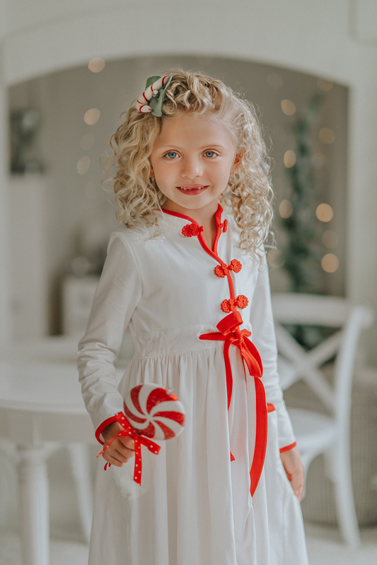 A young girl is wearing a white robe kimono.  The buttery soft fabric is long sleeved with red piping trim.  There are toggle buttons and a red ribbon tie at the waist. A perfect lounge piece to watch a White Christmas. 