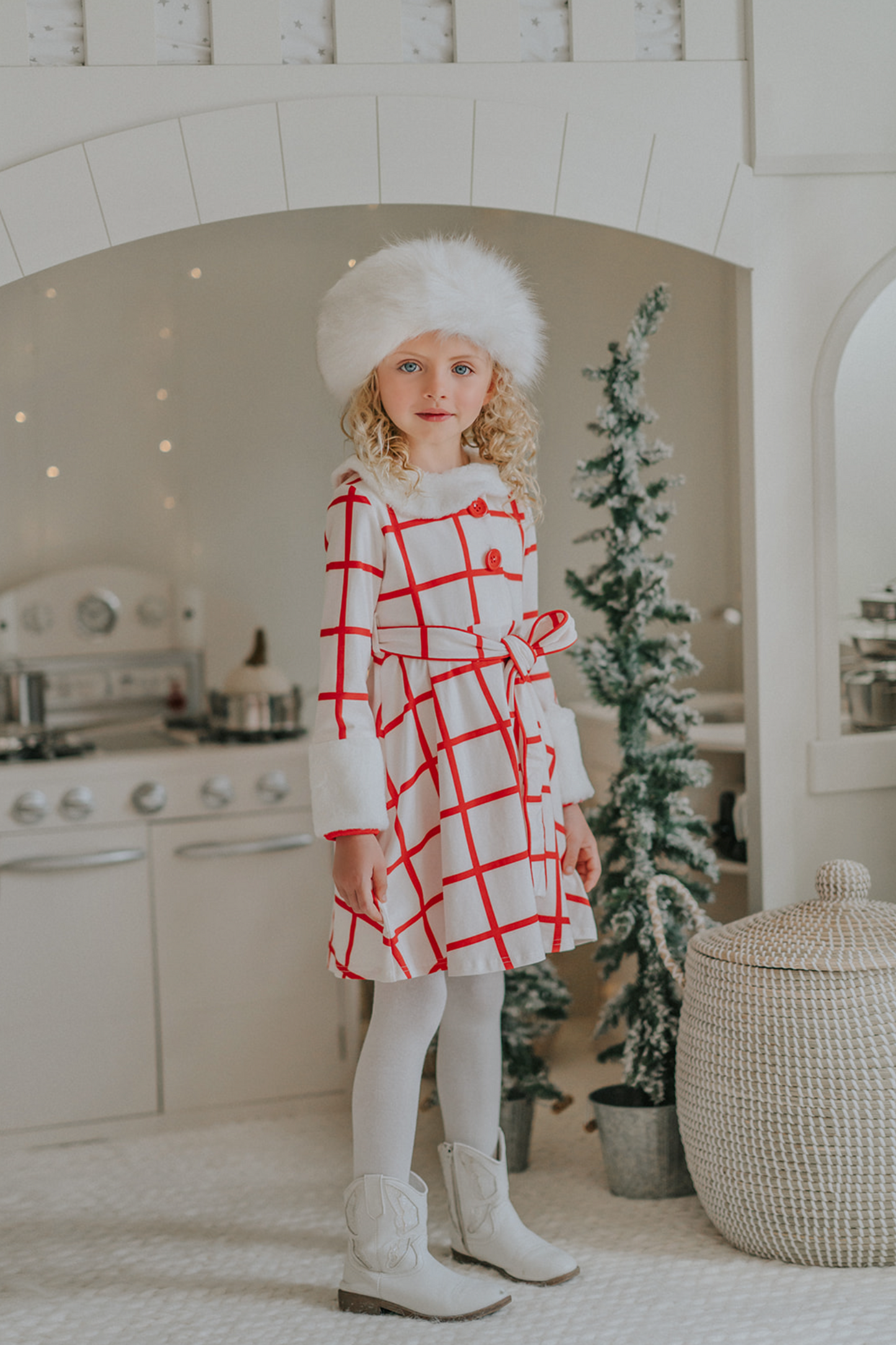 A young girl wears a white and red plaid windowpane dress.  The dress has red buttons at the top and a belt tie.  The collar and cuffs are a soft white fur. 