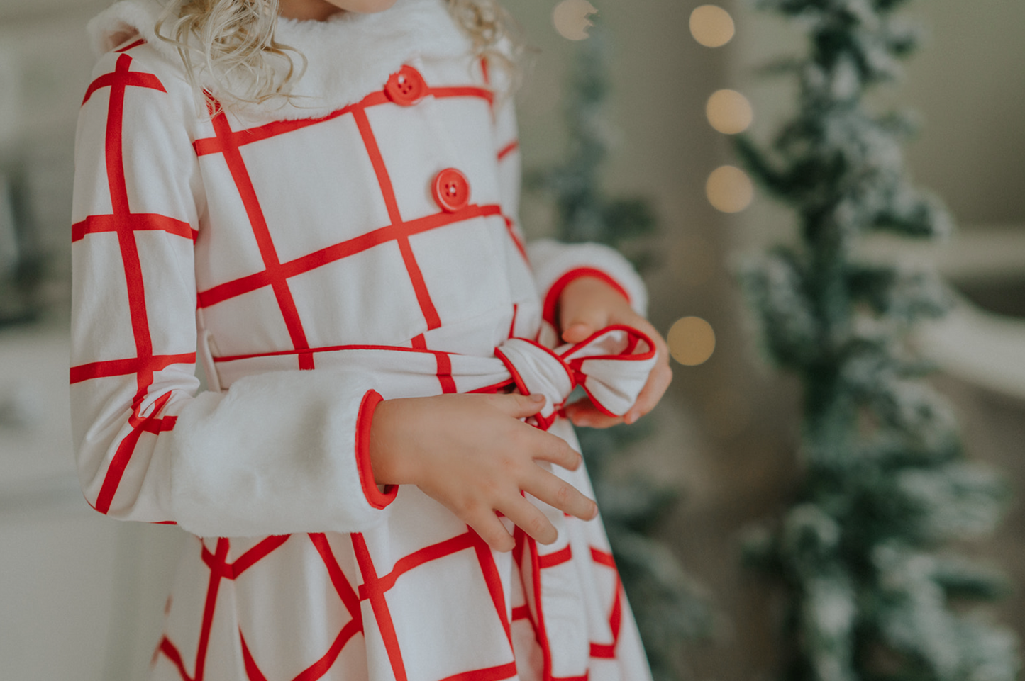 A young girl wears a white and red plaid windowpane dress.  The dress has red buttons at the top and a belt tie.  The collar and cuffs are a soft white fur. 