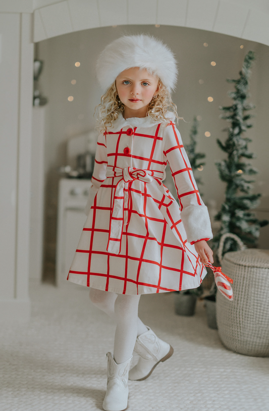 A young girl wears a white and red plaid windowpane dress.  The dress has red buttons at the top and a belt tie.  The collar and cuffs are a soft white fur. 