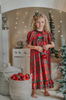 A young girl is wearing The Original Clara Nutcracker Gown in Red Plaid, featuring red plaid ruffles and a green bow. The plaid has white, green and black with red background. The buttery soft fabric is cozy and sensory friendly.  The puff sleeves with ribbon detail and the ruffles at the hem add a traditional classic look. 