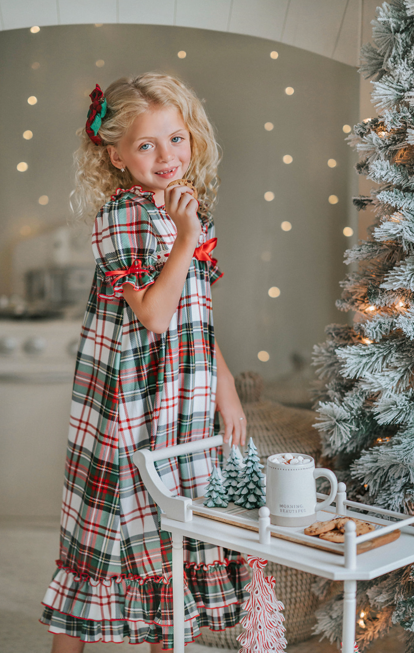 A young girl with curly blonde hair stands in front of a beautifully decorated Christmas tree. She is wearing "The Original Clara Nutcracker Gown in White Plaid" and smiling radiantly. Next to her, there's a cart brimming with holiday decorations, including small Christmas trees and a glowing candle. The background features soft, warm lighting that enhances the festive ambiance.