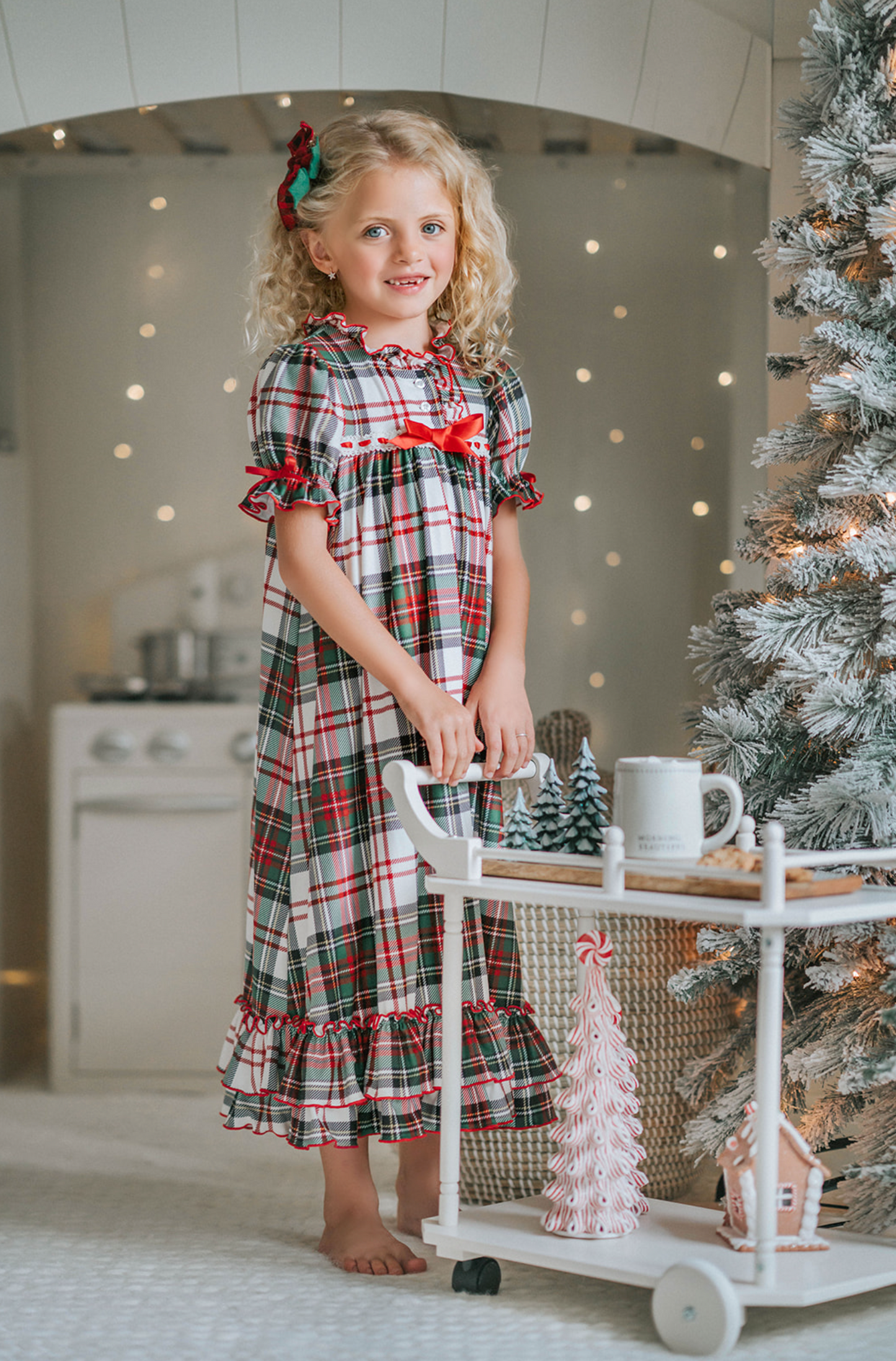 A young girl with curly blonde hair stands in front of a beautifully decorated Christmas tree. She is wearing "The Original Clara Nutcracker Gown in White Plaid" and smiling radiantly. Next to her, there's a cart brimming with holiday decorations, including small Christmas trees and a glowing candle. The background features soft, warm lighting that enhances the festive ambiance.