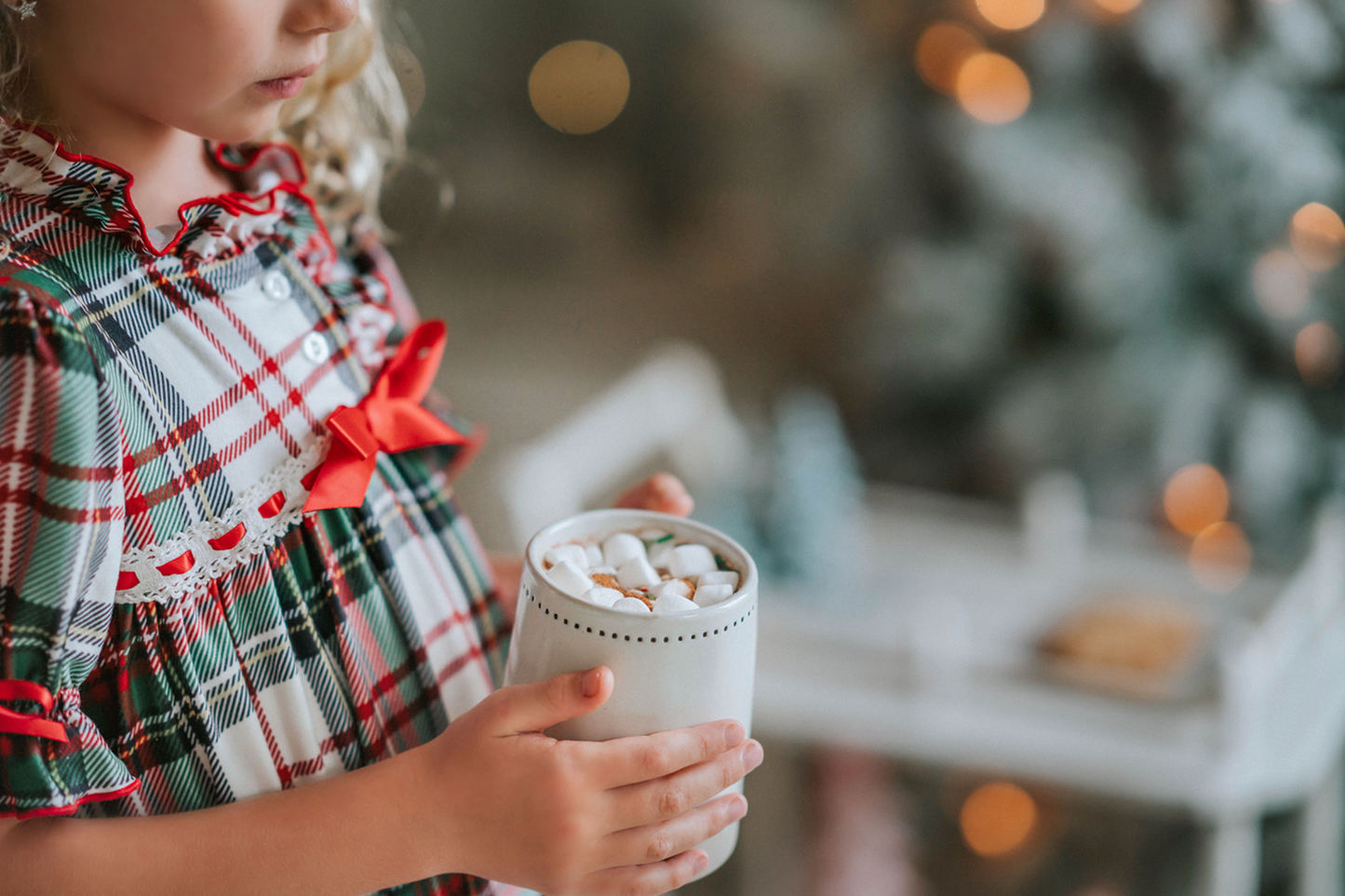 A young girl with curly blonde hair stands in front of a beautifully decorated Christmas tree. She is wearing "The Original Clara Nutcracker Gown in White Plaid" and smiling radiantly. Next to her, there's a cart brimming with holiday decorations, including small Christmas trees and a glowing candle. The background features soft, warm lighting that enhances the festive ambiance.