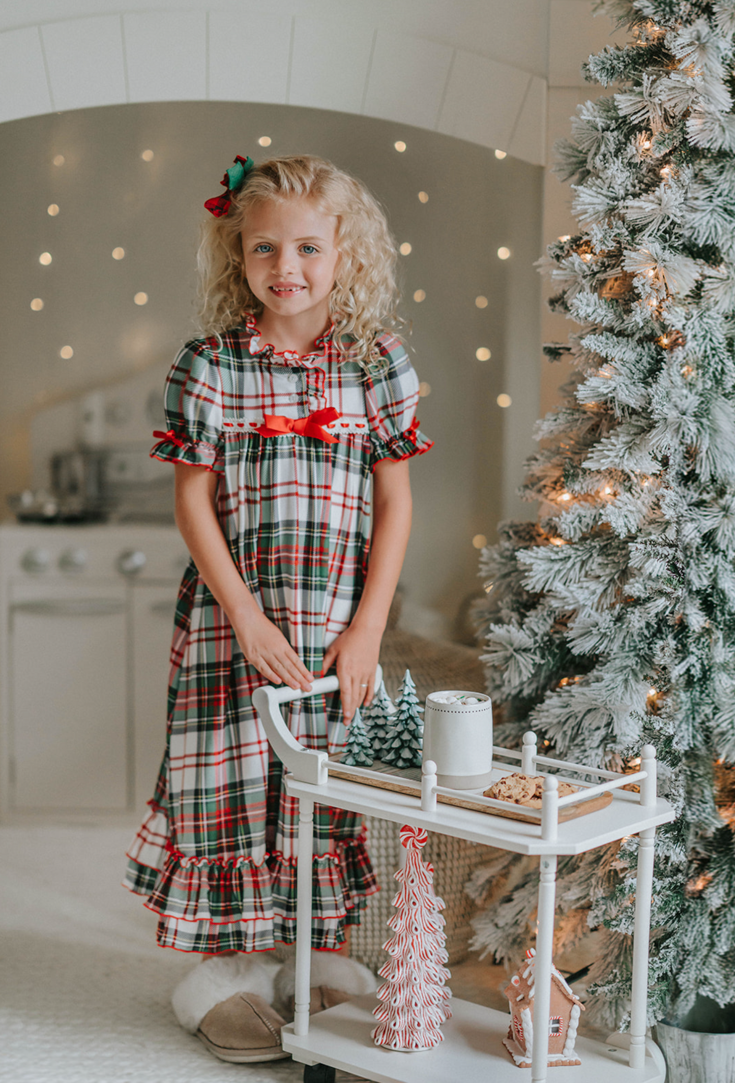 A young girl is wearing The Original Clara Nutcracker Gown in White Plaid, featuring white plaid ruffles and a bright red bow. The buttery soft fabric is cozy and sensory friendly.  The puff sleeves with ribbon detail and the ruffles at the hem add a traditional classic look. 