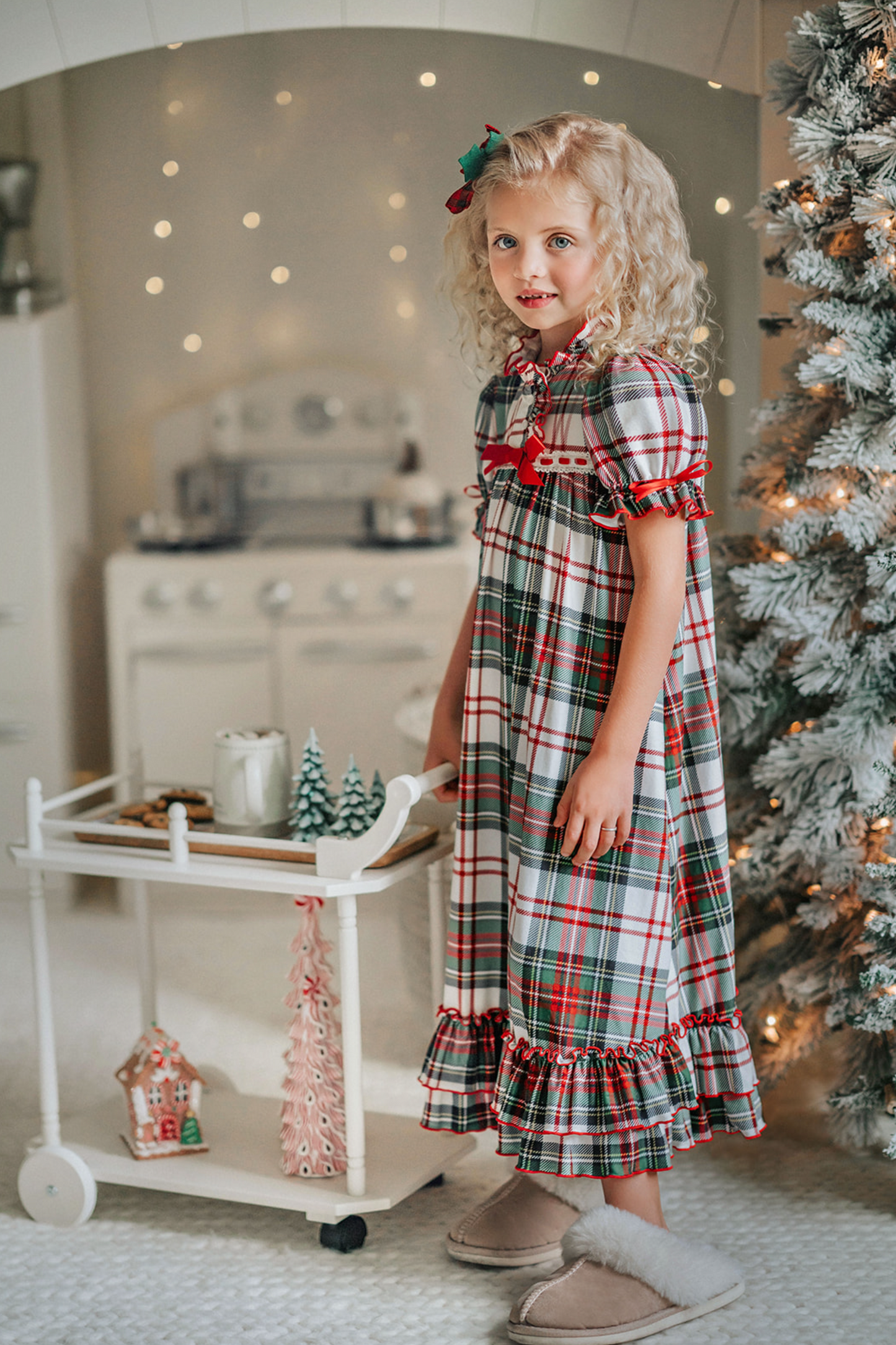 A young girl with curly blonde hair stands in front of a beautifully decorated Christmas tree. She is wearing "The Original Clara Nutcracker Gown in White Plaid" and smiling radiantly. Next to her, there's a cart brimming with holiday decorations, including small Christmas trees and a glowing candle. The background features soft, warm lighting that enhances the festive ambiance.