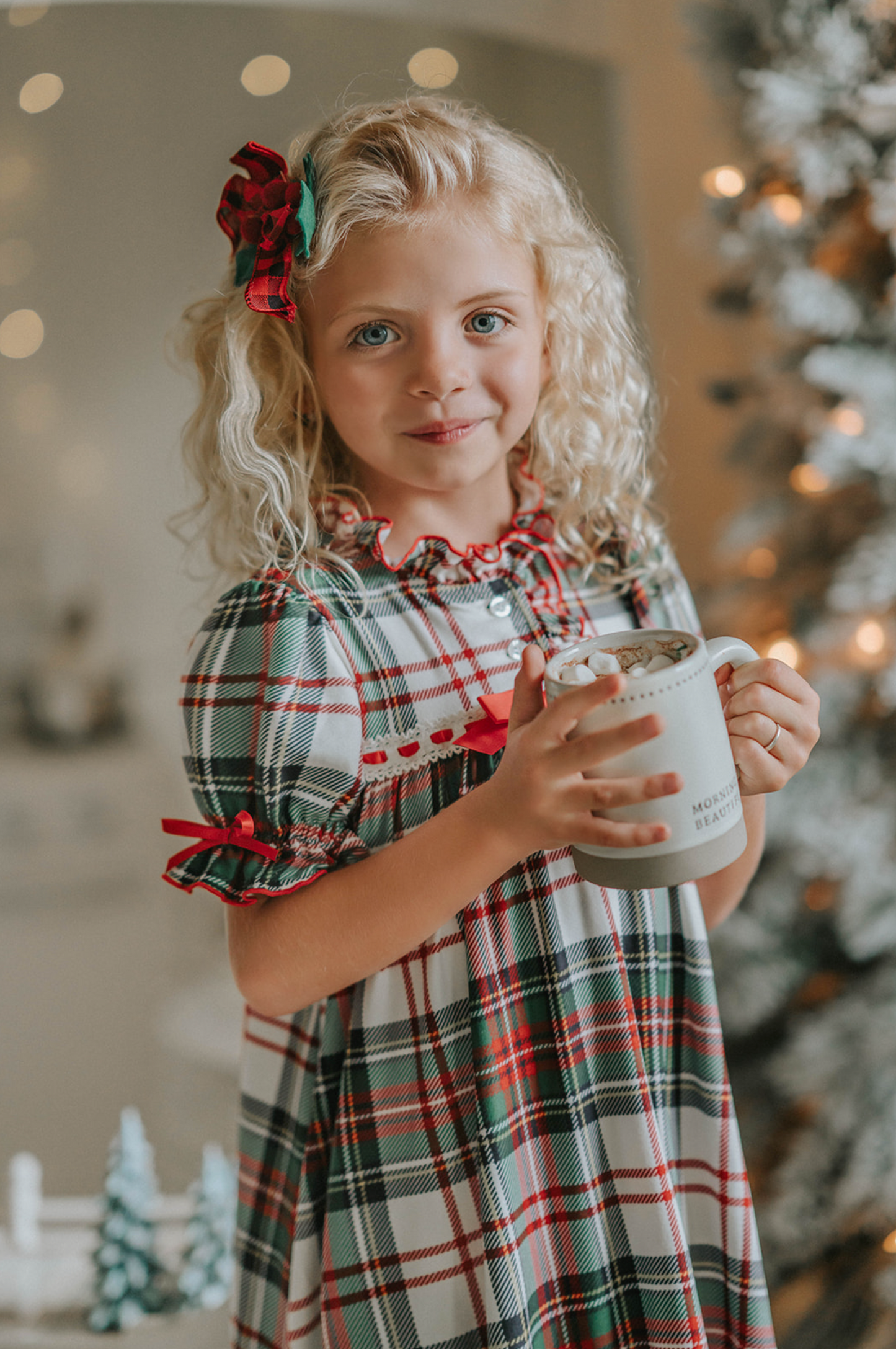 A young girl with curly blonde hair stands in front of a beautifully decorated Christmas tree. She is wearing "The Original Clara Nutcracker Gown in White Plaid" and smiling radiantly. Next to her, there's a cart brimming with holiday decorations, including small Christmas trees and a glowing candle. The background features soft, warm lighting that enhances the festive ambiance.